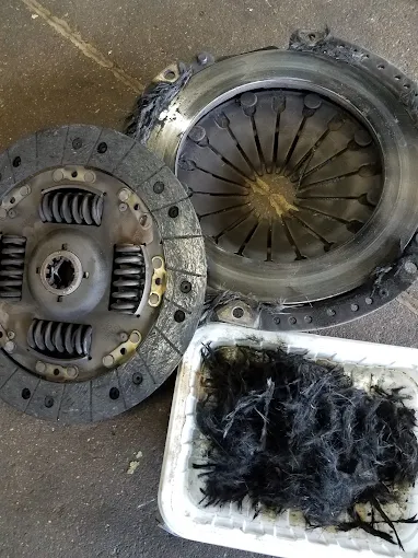 A worn-out clutch friction disc from a customer's vehicle on the shop floor, with the shredded lining material collected in a tray beside it