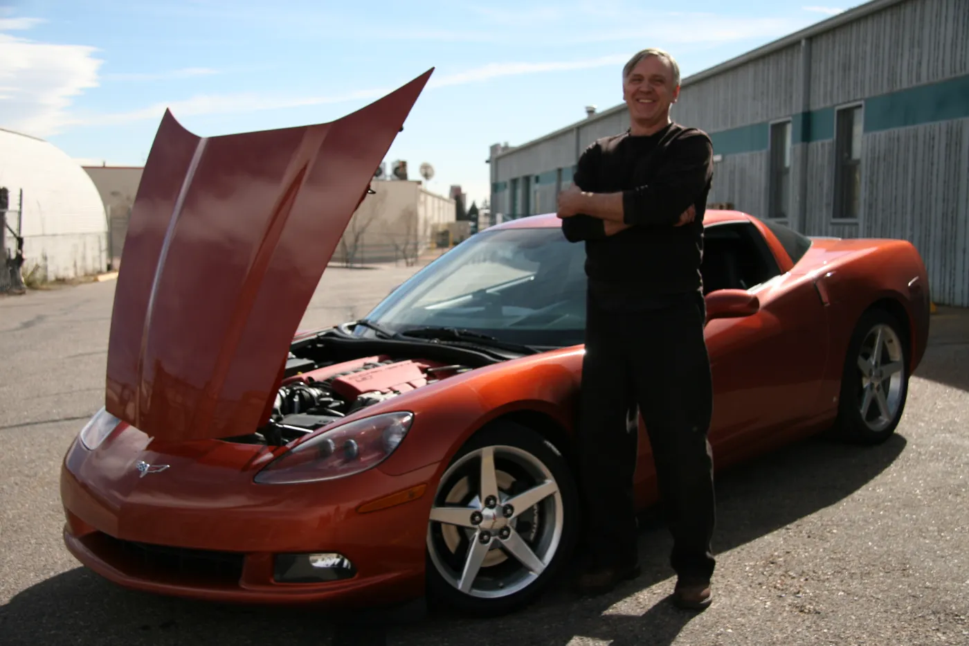 Ed standing with folded arms beside an orange Chevrolet Corvette with its hood raised, outside the shop