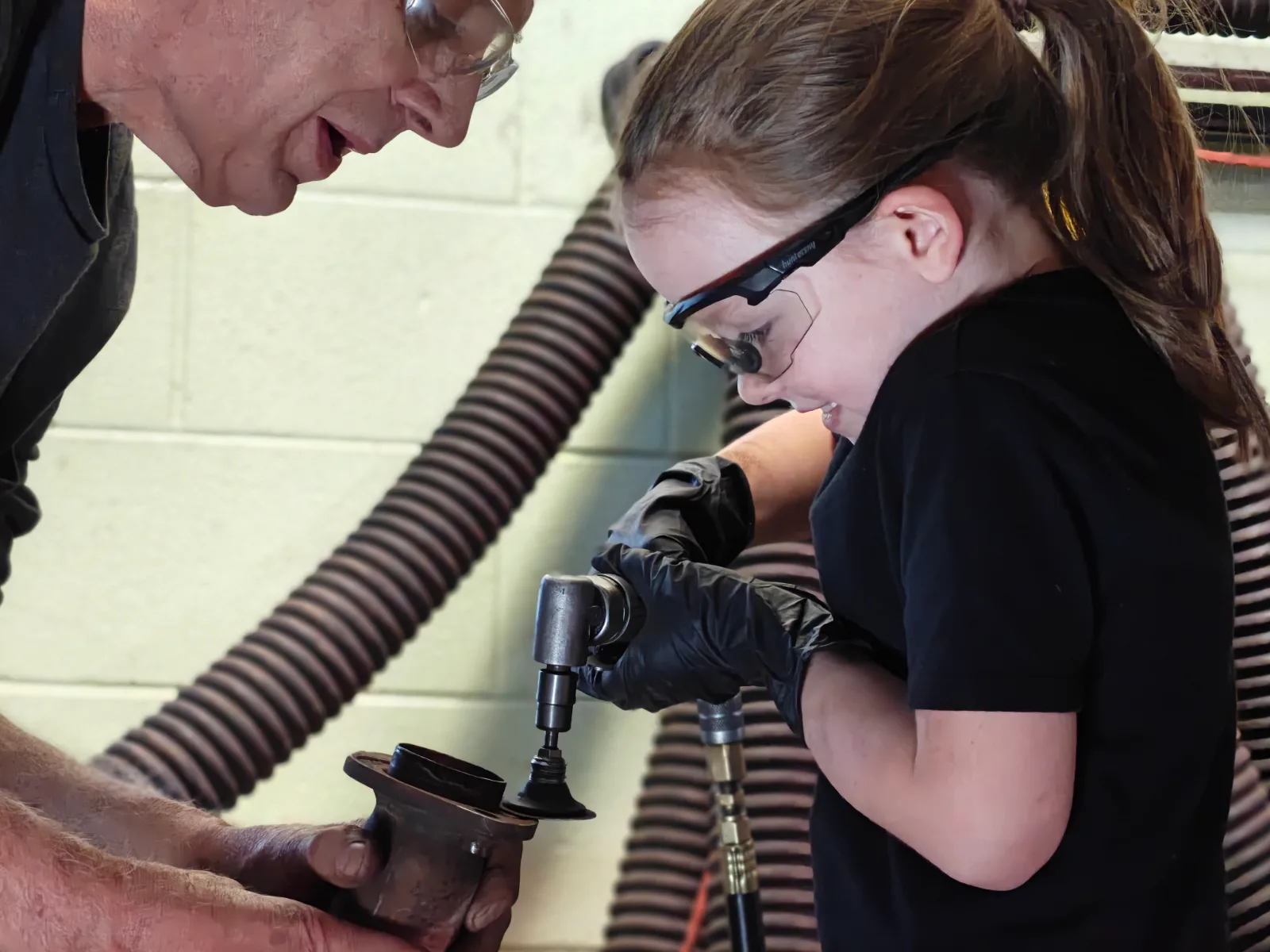 Ed steadying a component on the bench in safety glasses while his young granddaughter, also in safety glasses, works on it with an air tool