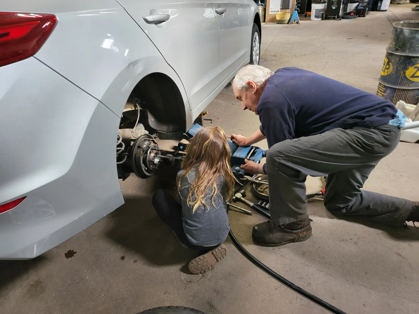 Ed kneeling at the exposed rear hub of a silver sedan with his granddaughter crouched beside him, both working on the assembly