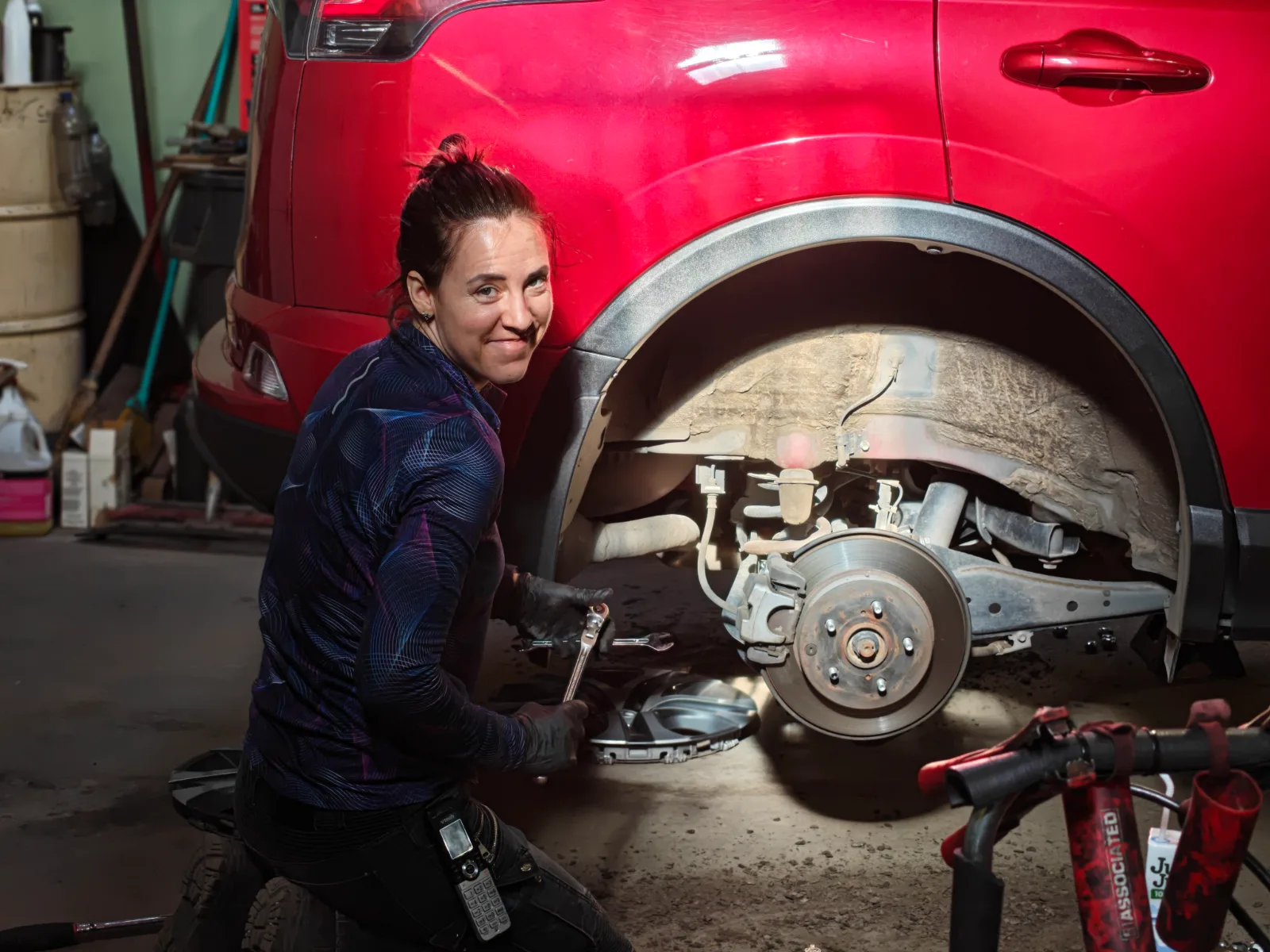 Nicole kneeling beside the exposed rear brake rotor of a red SUV, wrench in hand, looking up at the camera