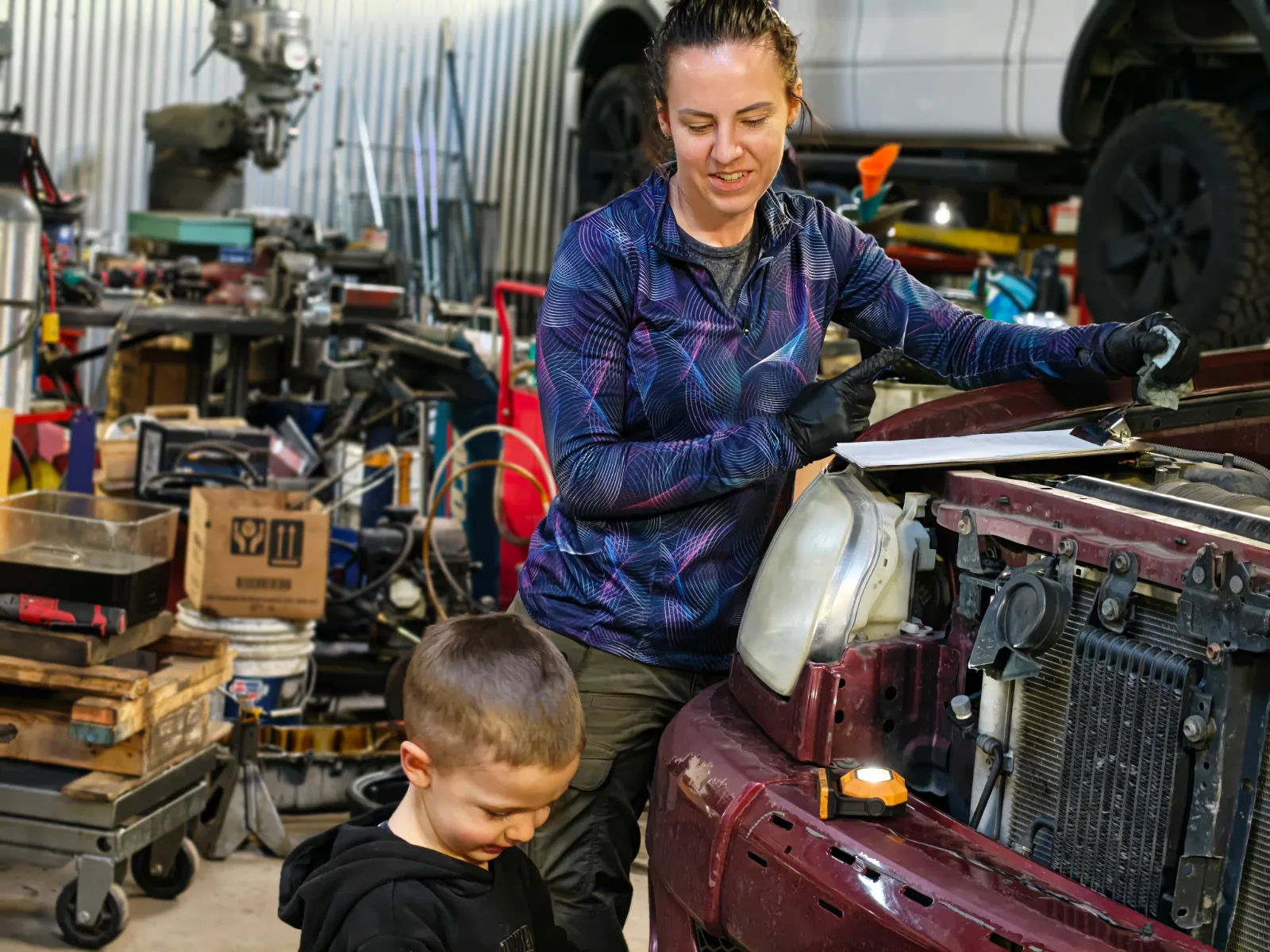 Nicole working under the raised hood of a maroon truck with the headlight and radiator support exposed, a young boy standing beside her