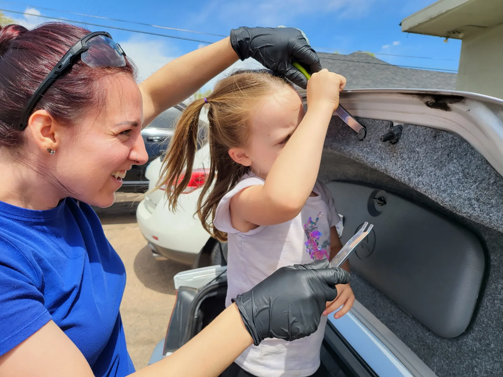 Nicole laughing with a small girl in the shop yard, the child holding trim tools over an open car trunk